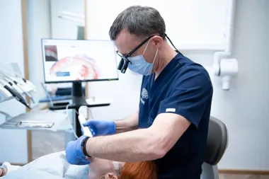 Dentysta w masce i rękawiczkach leczy pacjentkę na fotelu stomatologicznym.