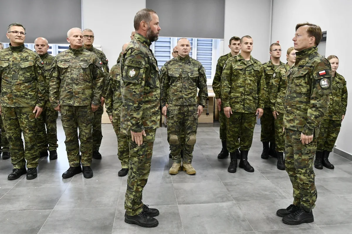 Grupa żołnierzy w mundurach kamuflażowych stoi w sali w formacji, dwaj oficerowie rozmawiają z przodu.