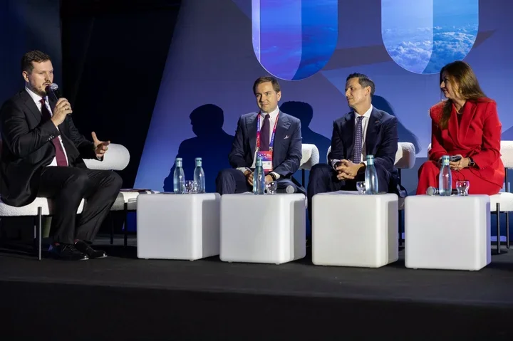 Cztery osoby ubrane biznesowo siedzą na scenie podczas panelu dyskusyjnego, jeden mężczyzna przemawia do mikrofonu.