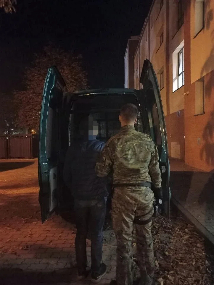 W nocy funkcjonariusz w mundurze wojskowym prowadzi mężczyznę do zielonego furgonu przy budynku.