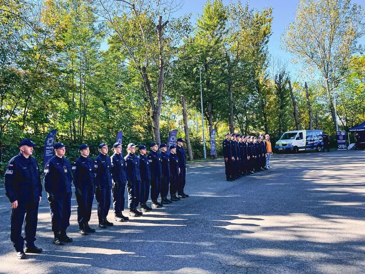 Funkcjonariusze policji w granatowych mundurach stoją w szeregu na asfaltowej powierzchni w otoczeniu drzew, w tle pojazd służbowy i namiot promocyjny.