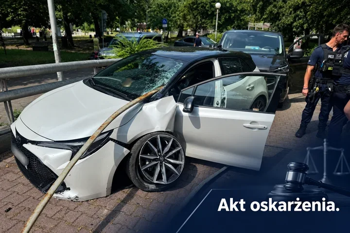 Biały samochód osobowy z uszkodzoną karoserią i rozbitym przednią szybą po zderzeniu z barierą stoi na brukowanej nawierzchni, obok interweniują policjanci.