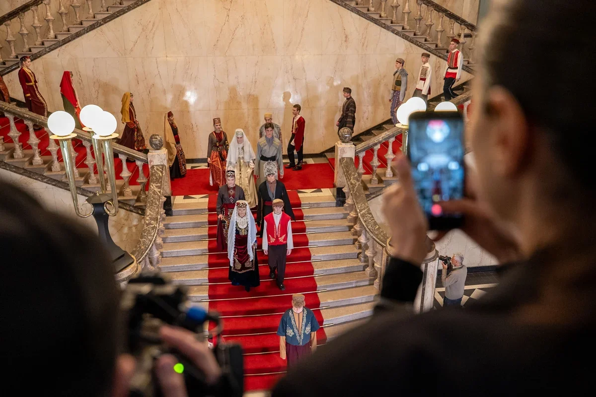 Modele w strojach ludowych prezentują się na monumentalnym marmurowym schodzie z czerwonym dywanem, a widzowie fotografują pokaz telefonami.