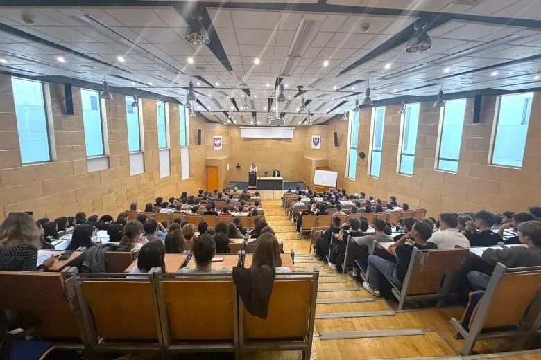 Widok z tyłu na pełną aulę wykładową z drewnianymi ławkami, gdzie słuchacze siedzą przed sceną z dwoma osobami za pulpitem.