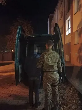 W nocy funkcjonariusz w mundurze wojskowym prowadzi mężczyznę do zielonego furgonu przy budynku.