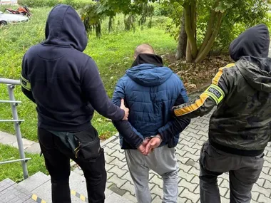 Funkcjonariusze policji w mundurach i bluzach z kapturem zatrzymują mężczyznę w niebieskiej kurtce, który ma skrępowane ręce kajdankami.
