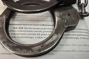 Metalowe kajdanki leżą na otwartym kodeksie prawnym, a w okienku jednej z nich widać fragment artykułu 244 o niestosowaniu się do orzeczonych środków karnych.