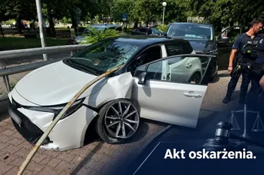 Biały samochód osobowy z uszkodzoną karoserią i rozbitym przednią szybą po zderzeniu z barierą stoi na brukowanej nawierzchni, obok interweniują policjanci.