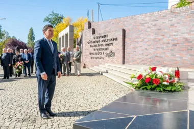 Mężczyzna w garniturze stoi przed pomnikiem z czerwonej cegły podczas uroczystości z wieniecami i uczestnikami w tle.
