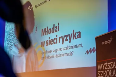 Fragment sali konferencyjnej z ekranem wyświetlającym hasło konferencji „Młodzi w sieci ryzyka” oraz pulpitem z logo WYŻSZEJ SZKOŁY.