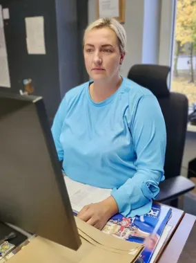Kobieta w niebieskiej bluzce siedzi przy biurku przed monitorem komputera w pomieszczeniu biurowym.