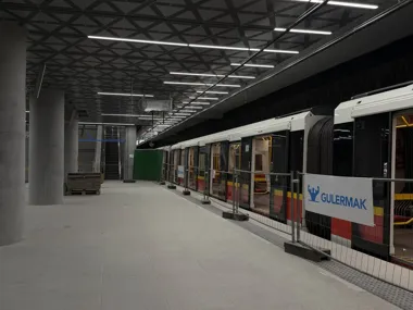 Pociąg metra stoi na nowoczesnej, jeszcze nieoddanej do użytku stacji podziemnej, przy peronie widoczne są elementy wykończeniowe i ogrodzenie budowlane.