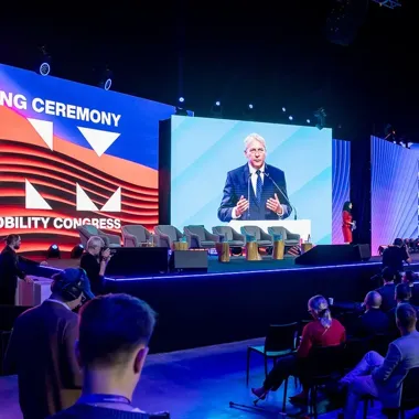 Mężczyzna w garniturze przemawia z mównicy podczas uroczystego otwarcia kongresu mobilności, na scenie z fotelami i dużymi ekranami, widownia słucha w sali konf