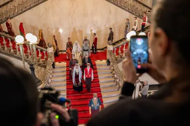 Modele w strojach ludowych prezentują się na monumentalnym marmurowym schodzie z czerwonym dywanem, a widzowie fotografują pokaz telefonami.