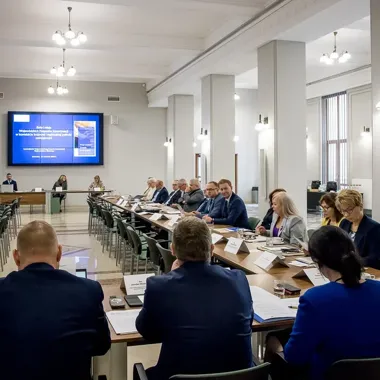 Uczestnicy konferencji lub posiedzenia siedzą przy długich stołach w sali konferencyjnej, na ekranie wyświetlana jest prezentacja dotycząca wojewódzkich zespołó