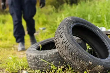 Zużyte opony leżą na trawie, w tle widać nogi przechodzącej osoby w ciemnych spodniach i kaloszach.