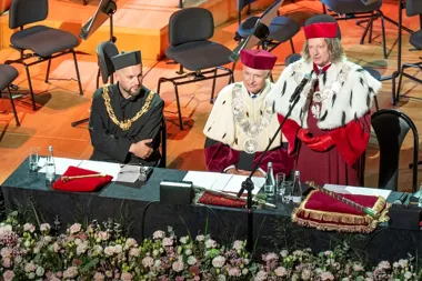 Trzej mężczyźni w uroczystych, historycznych strojach akademickich z insygniami zasiadają przy stole na scenie, jeden z nich przemawia do mikrofonu podczas ofic