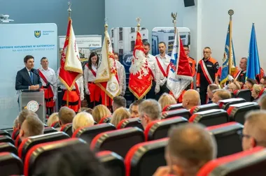 Mężczyzna przemawia z mównicy podczas uroczystości z udziałem pocztów sztandarowych i zaproszonych gości.