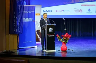 Mężczyzna w garniturze przemawia z mównicy na scenie podczas konferencji o bezpieczeństwie seniorów, w tle banery CBZC, NASK i innych partnerów.