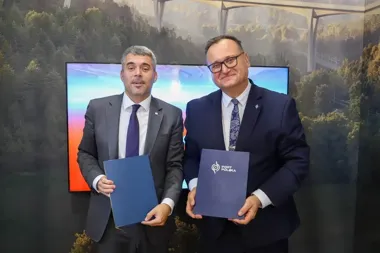 Dwaj mężczyźni w garniturach trzymają podpisane dokumenty podczas uroczystości, w tle ekran i fotografia mostu.