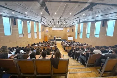 Widok z tyłu na pełną aulę wykładową z drewnianymi ławkami, gdzie słuchacze siedzą przed sceną z dwoma osobami za pulpitem.