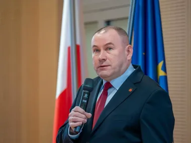 Mężczyzna w garniturze z mikrofonem przemawia podczas oficjalnego wydarzenia, w tle flagi Polski i Unii Europejskiej.