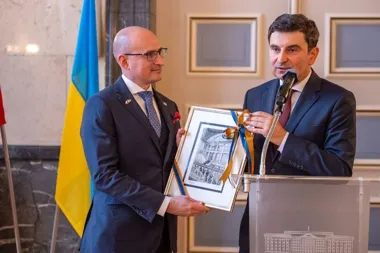 Dwaj mężczyźni w garniturach podczas uroczystości w sali z flagą Ukrainy w tle, jeden wręcza drugiemu oprawiony rysunek związany wstążką w barwach ukraińskich.