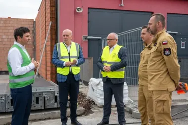 Czterech mężczyzn, w tym dwaj w mundurach i trzej w kamizelkach odblaskowych, stoi na terenie budowy przed czerwoną halą przemysłową i rozmawia.