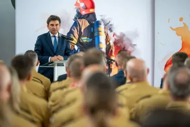 Mężczyzna w garniturze przemawia z mównicy do grupy słuchaczy w mundurach, za nim plakat z wizerunkiem strażaka.