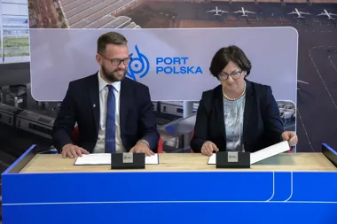Dwie osoby w strojach biznesowych siedzą przy niebieskim stole i podpisują dokumenty podczas wydarzenia związanego z Portem Polska, na tle grafiki przedstawiają
